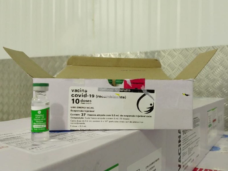 Do lado esquerdo há um frasco da vacina de Oxford com rótulo verde. Atrás aparece uma caixa branca aberta, onde estão as demais ampolas, com um rótulo onde está escrito vacina covid-19 (recombinante) e demais informações sobre o imunizante